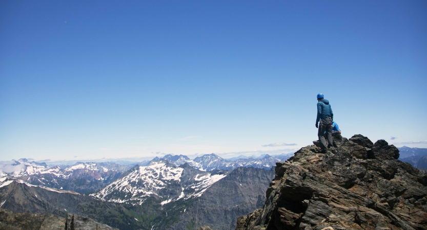 Two people stand on a rocky cliff at very high elevation, looking out over the vast mountainous and snowy landscape below.